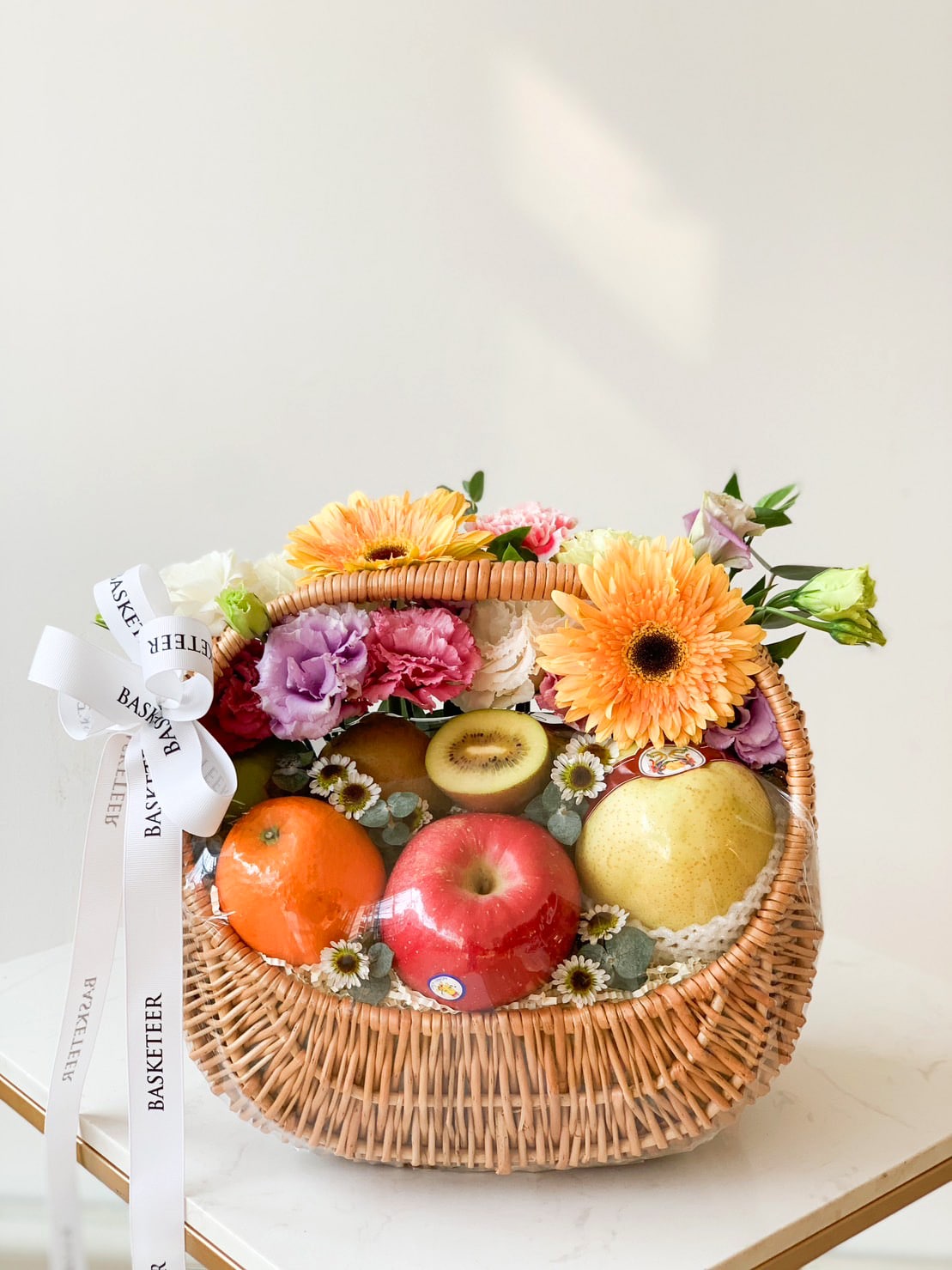 A Luscious Fruit Harvest Basket filled with assorted fruits, including an apple, oranges, a kiwi, and a melon, is adorned with a variety of colorful flowers such as gerberas, carnations, and daisies. A white ribbon with the text 