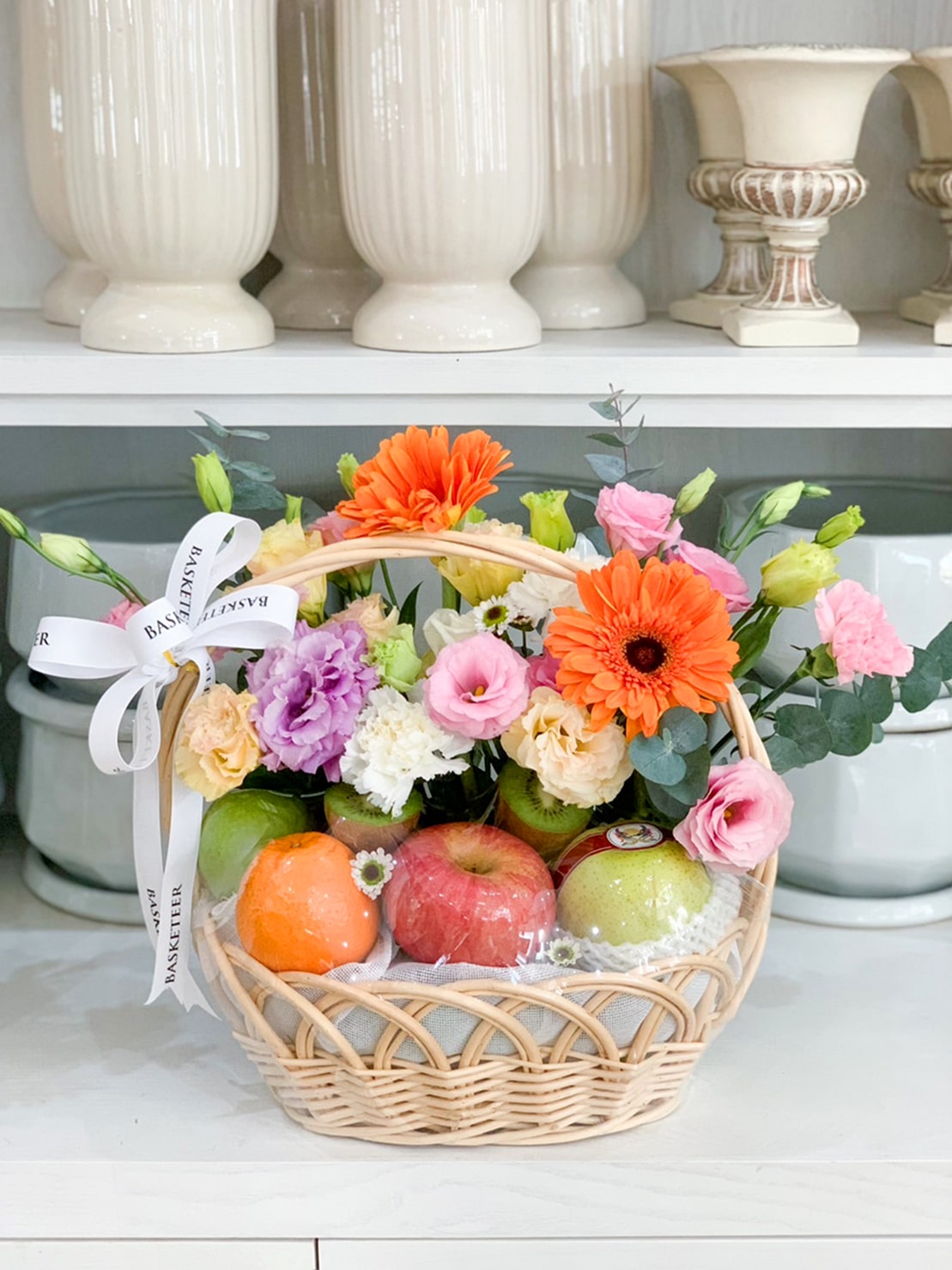 A luscious fruit harvest basket brimming with various flowers, including orange gerberas, and a mix of purple, pink, and white blooms. The wicker basket also contains apples and an orange, adorned with a white ribbon featuring black text and set against a backdrop of white vases.