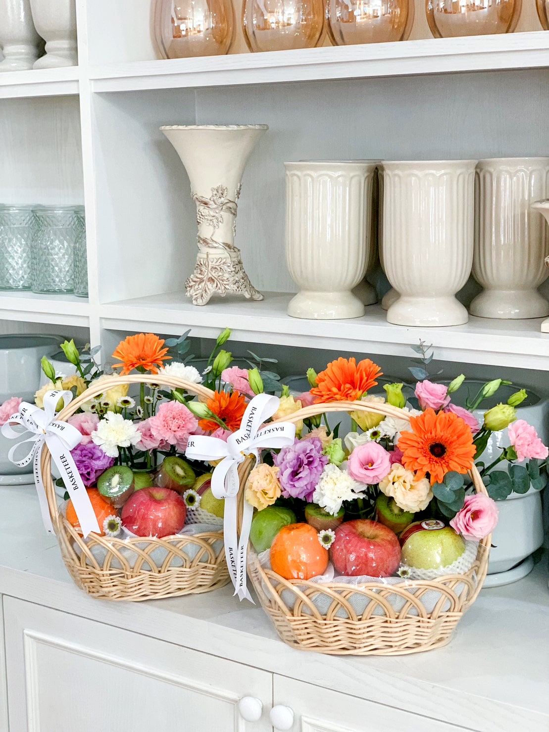 Two wicker Luscious Fruit Harvest Baskets, each adorned with colorful fresh flowers such as gerbera daisies, roses, and carnations, sit on a white shelf. Each basket also contains an assortment of fruits including apples and oranges. Behind the baskets are ceramic vases and jars on display.