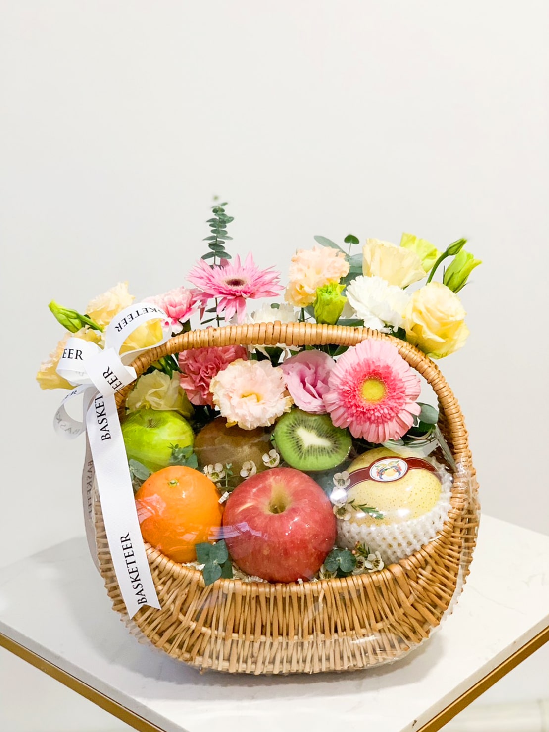 A wicker basket filled with an assortment of fresh fruits and flowers. The luscious Fruit Harvest Basket includes an apple, an orange, a kiwi, and a pear. The flowers feature various colorful blooms such as roses and gerbera daisies. The basket is adorned with a white ribbon.