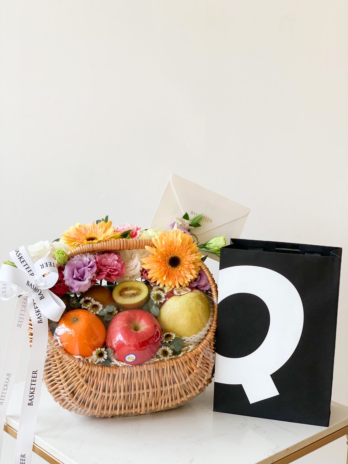 A luscious Fruit Harvest Basket filled with assorted fruits, including apples and citrus fruits, adorned with fresh flowers. A white envelope peeks out from the back of the basket. Next to it is a black gift bag with a large white letter 