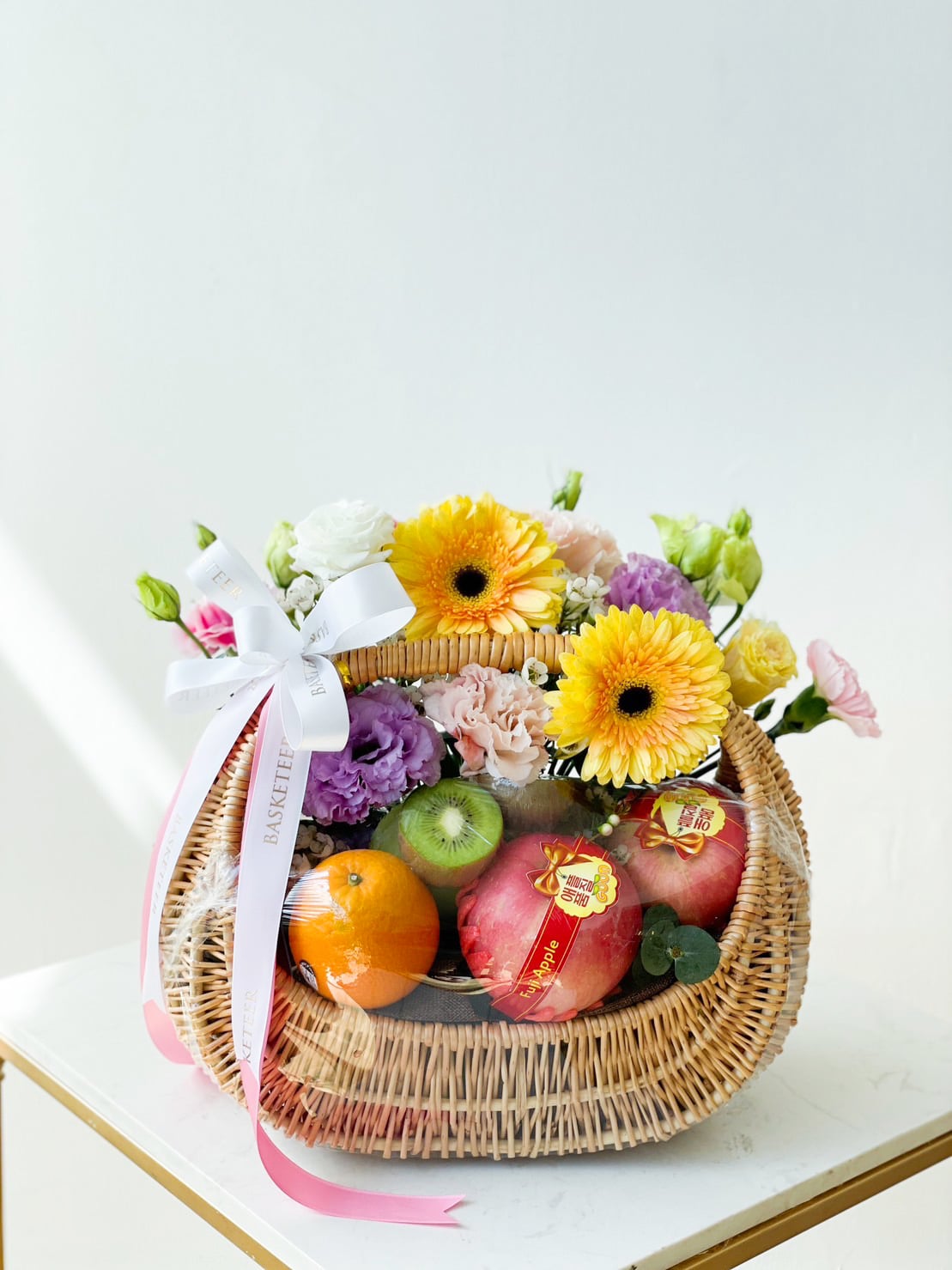 A wicker basket filled with assorted fruits, including apples and an orange, adorned with yellow, pink, and purple flowers. This Luscious Fruit Harvest Basket features a decorative white ribbon tied into a bow with 