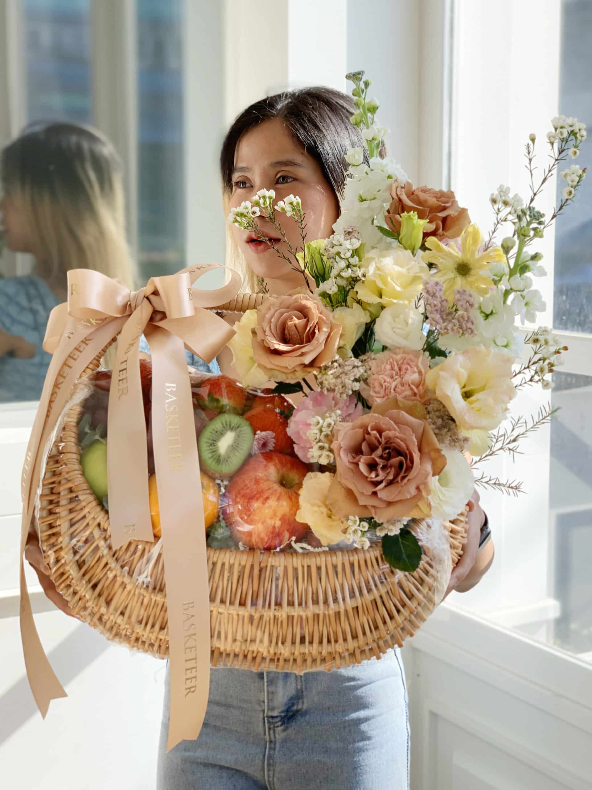 A person with long dark hair stands by a window, holding a large wicker basket. The basket contains fresh fruit like kiwi and apples, along with an arrangement of flowers, including roses and baby’s breath, tied with a ribbon labeled 