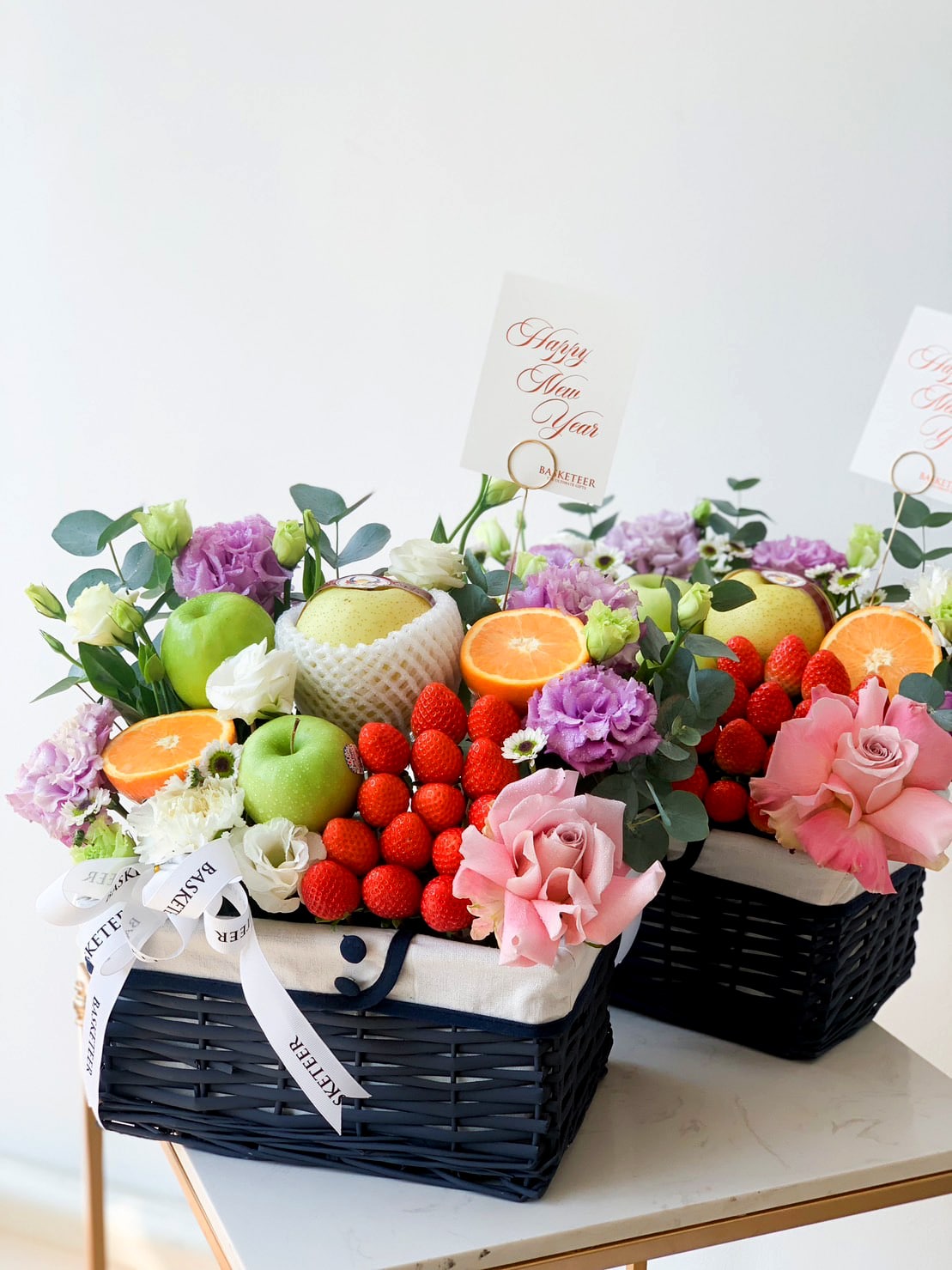 Two ornate gift baskets, brimming with an assortment of fruits like oranges, apples, pears, and strawberries, are adorned with roses and purple flowers. Each deluxe fruit basket comes with a 