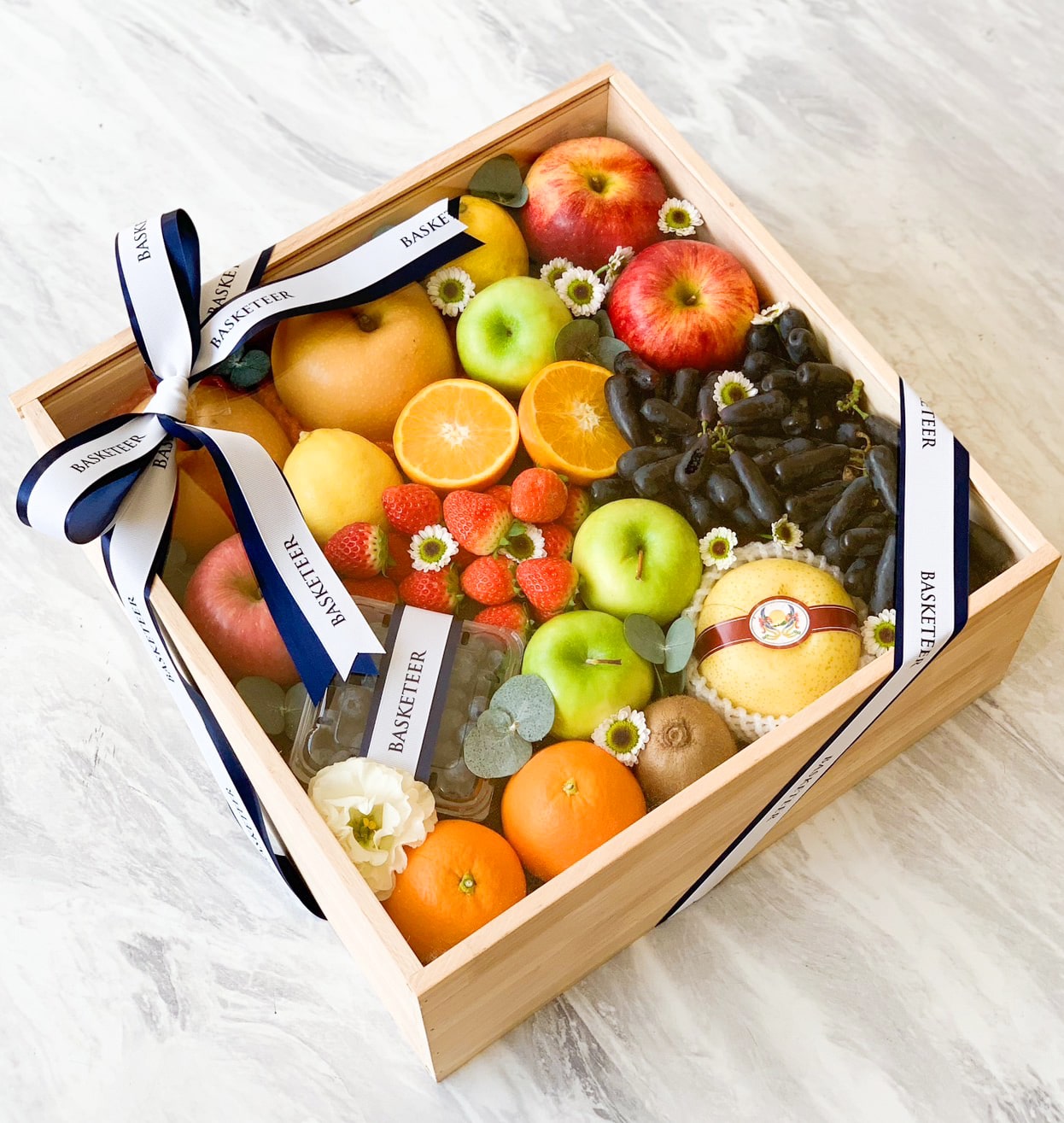 A wooden gift box filled with a bountiful variety of fresh fruits, including apples, oranges, strawberries, grapes, and citrus halves. The box exudes elegance with small white flowers and is wrapped with white ribbons adorned with the word 