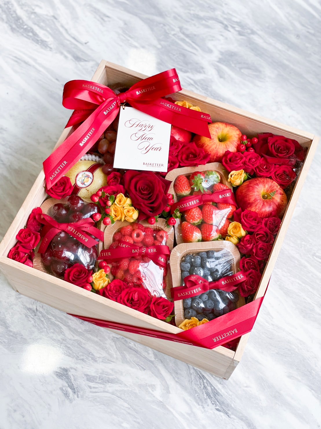 A wooden gift box filled with an assortment of fresh fruits, including apples, strawberries, cherries, and blueberries, alongside red and yellow roses. Elegantly arranged as a Mixed Berry Joy Gift, the box is adorned with red ribbons and a tag that says 