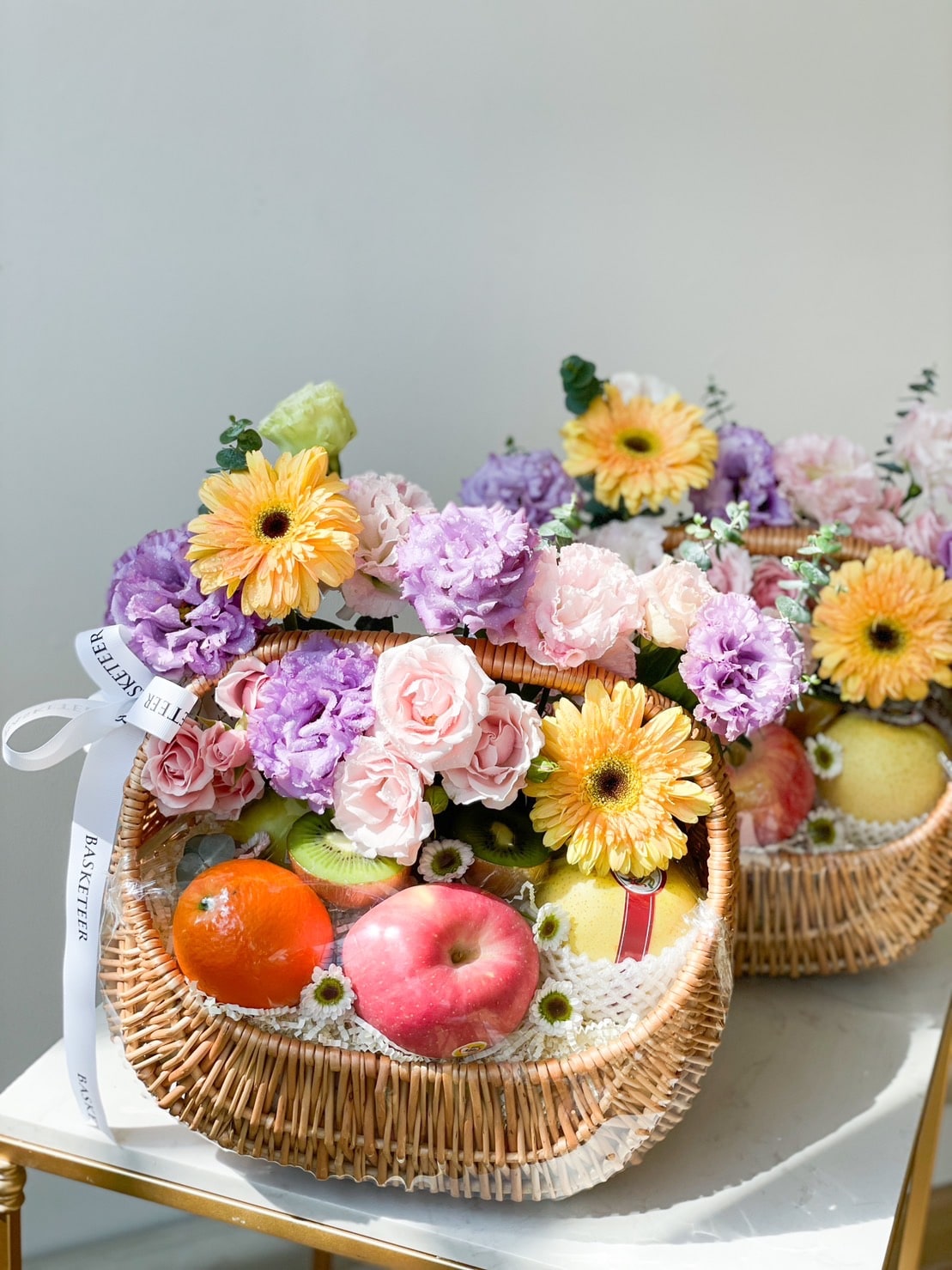 A wicker basket brimming with a colorful assortment of fresh flowers and fruits. The flowers include pink roses, purple carnations, and yellow daisies. The luscious fruit harvest basket features an apple, oranges, and pears. The basket is adorned with a white ribbon.