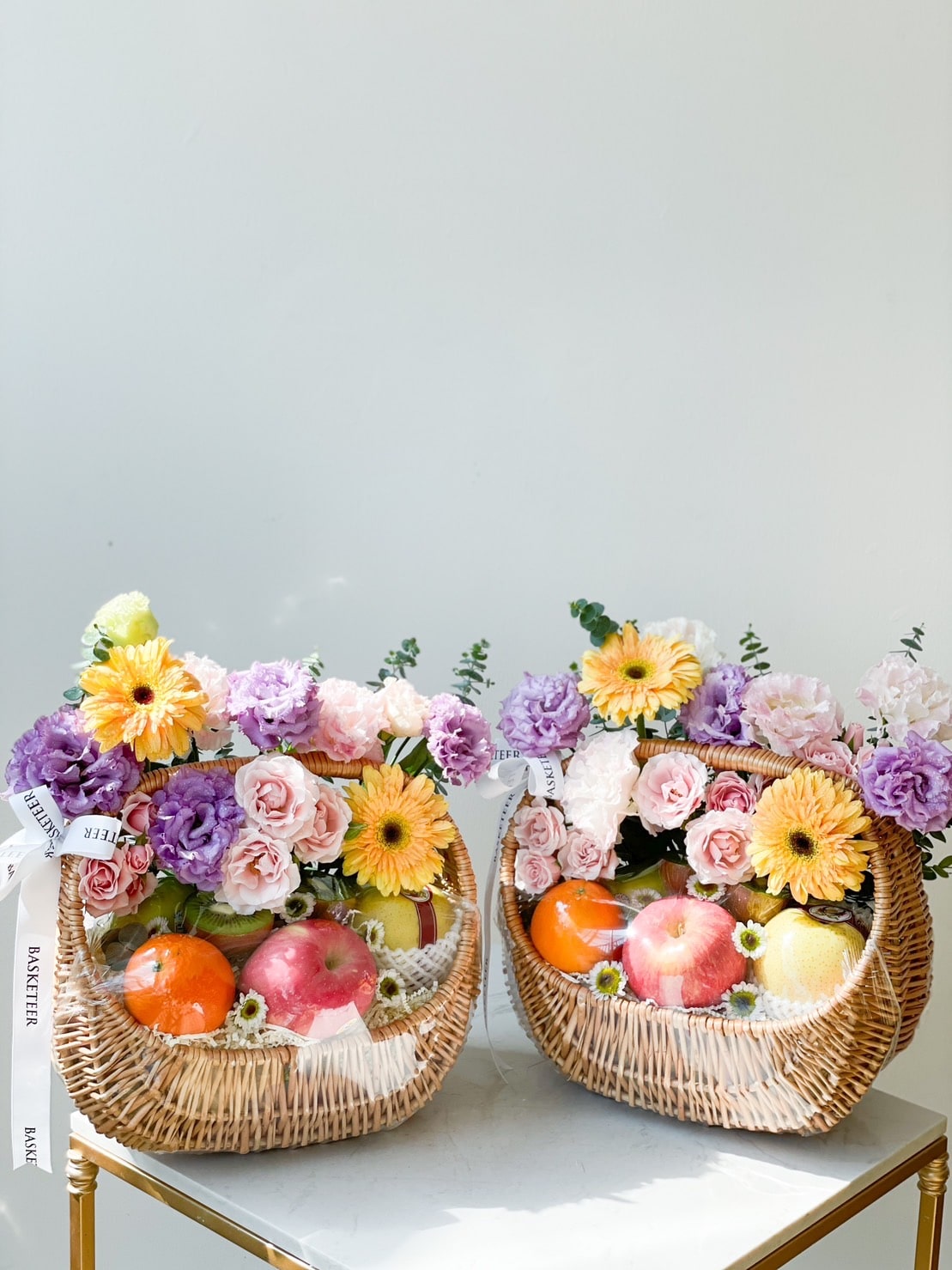 Two wicker baskets filled with an assortment of colorful flowers, including yellow and purple blooms, alongside a luscious fruit harvest of apples and oranges. Each basket is adorned with a white ribbon. They are placed on a white surface against a plain background.
