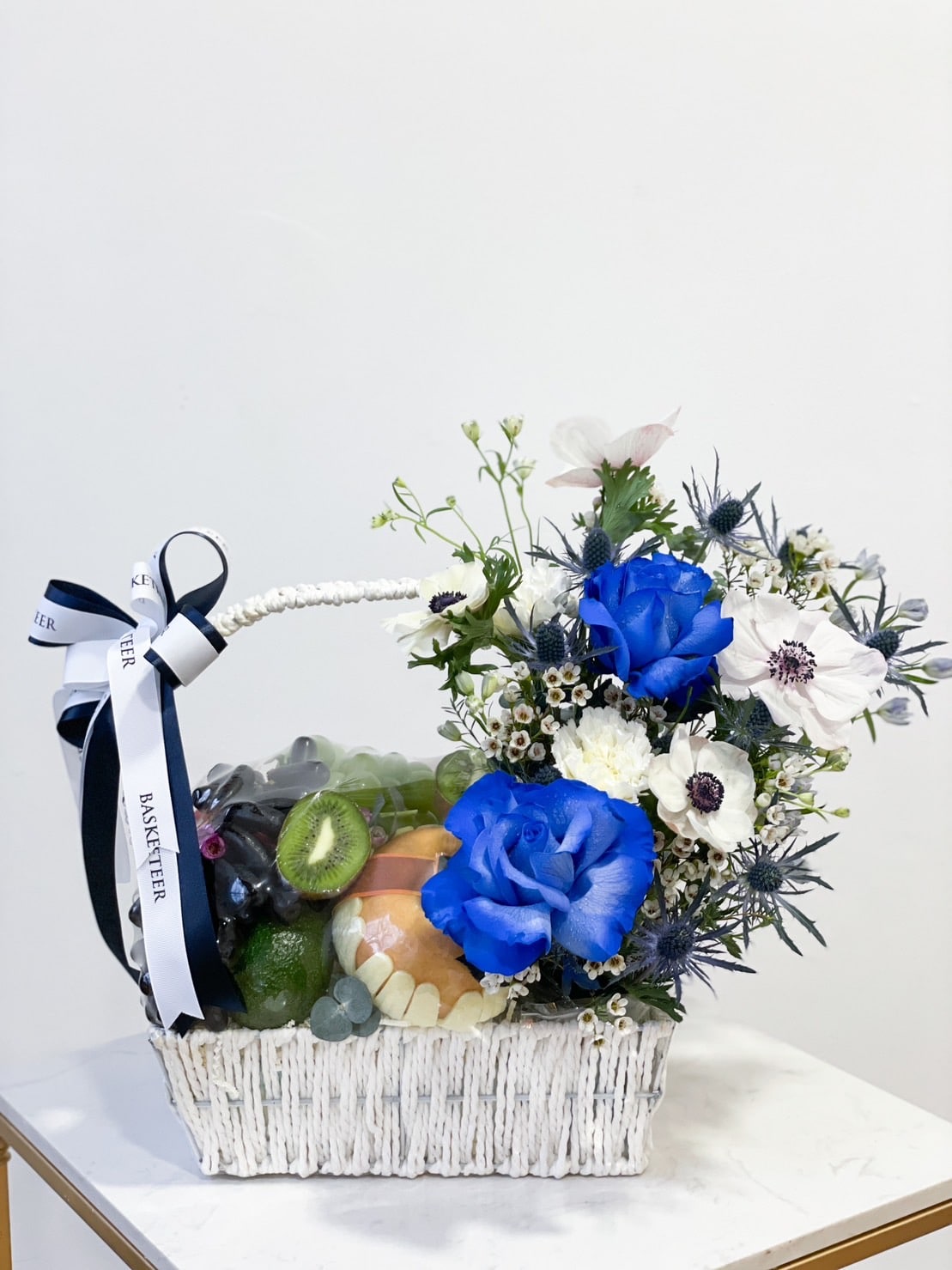The Blue Rose Fruit Basket is a white wicker basket adorned with blue roses and white flowers. It contains an assortment of fresh fruits, including kiwis, apples, grapes, and a green lime. The basket features a white ribbon with 