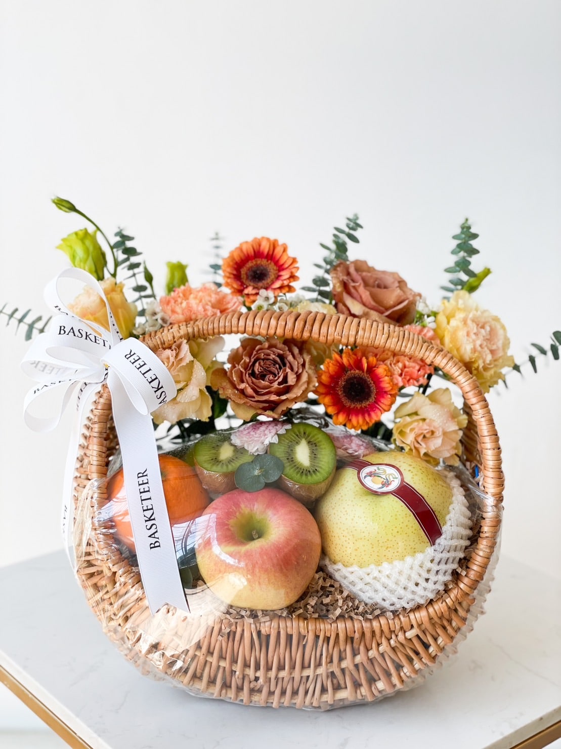A woven basket brimming with fresh fruits including apples, pears, kiwis, and an orange. Labeled 