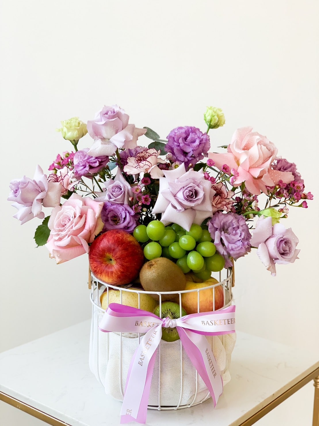 A decorative basket containing a variety of fresh fruits like apples, grapes, and pears is adorned with pastel-colored roses and carnations in shades of purple, pink, and green. The white base is tied with pink ribbons that say 