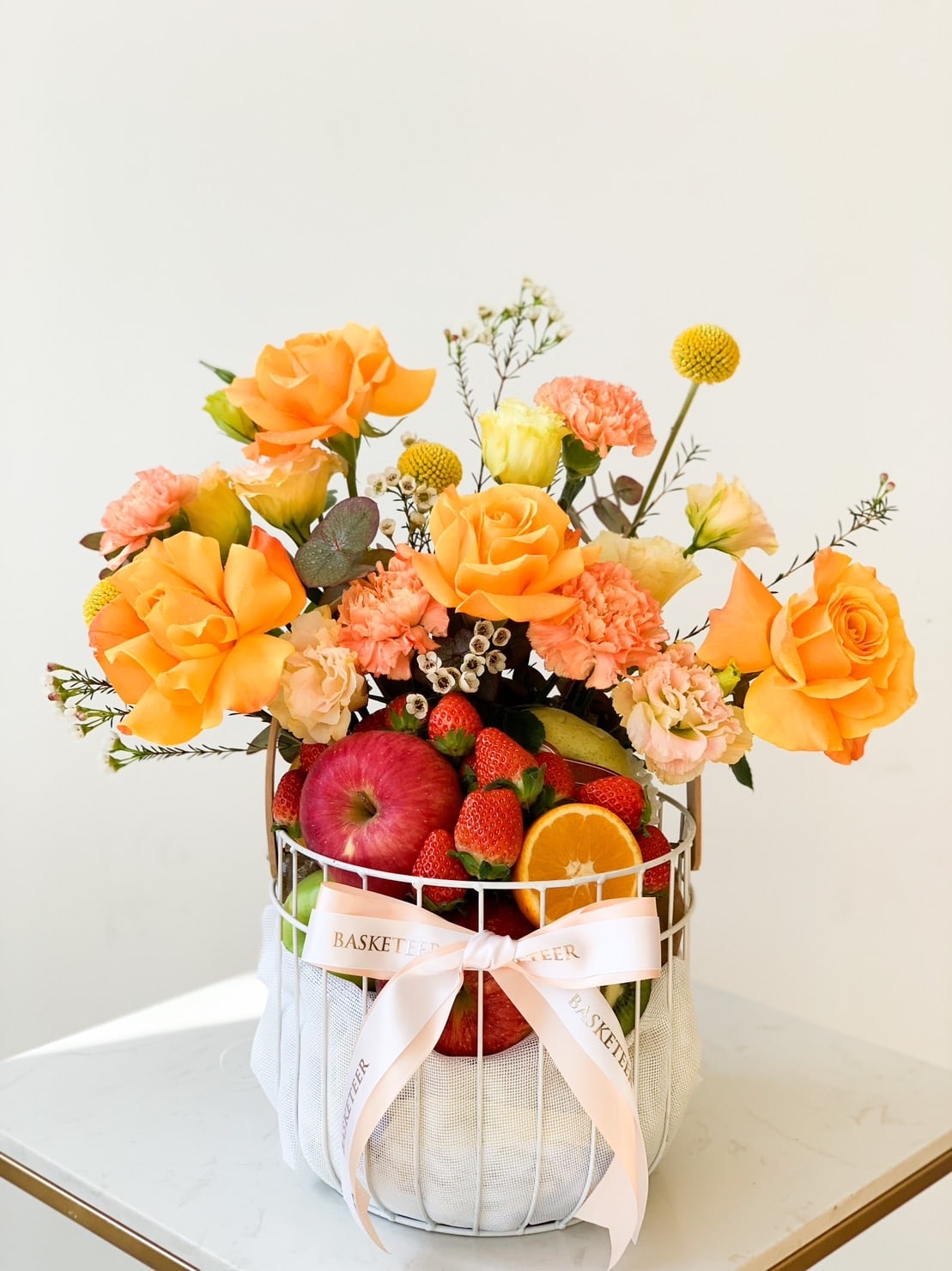 A round white basket containing a bouquet of orange roses, pink carnations, yellow billy balls, and various greenery. The Sunshine Fruits Basket also includes fresh fruits like apples and strawberries. It is adorned with a white ribbon that has 