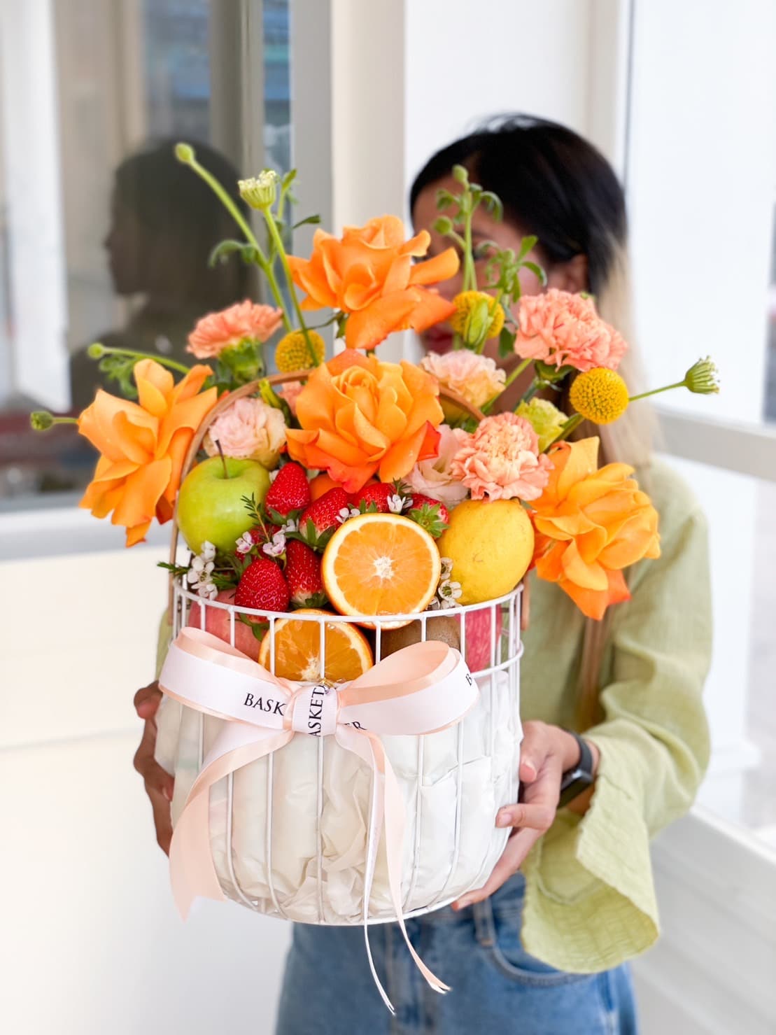 A person holds a white wire Sunshine Fruits Basket filled with an arrangement of bright, colorful flowers and various fruits, including oranges, apples, and strawberries. The basket is adorned with a white ribbon and set against a light-colored, softly-lit background.