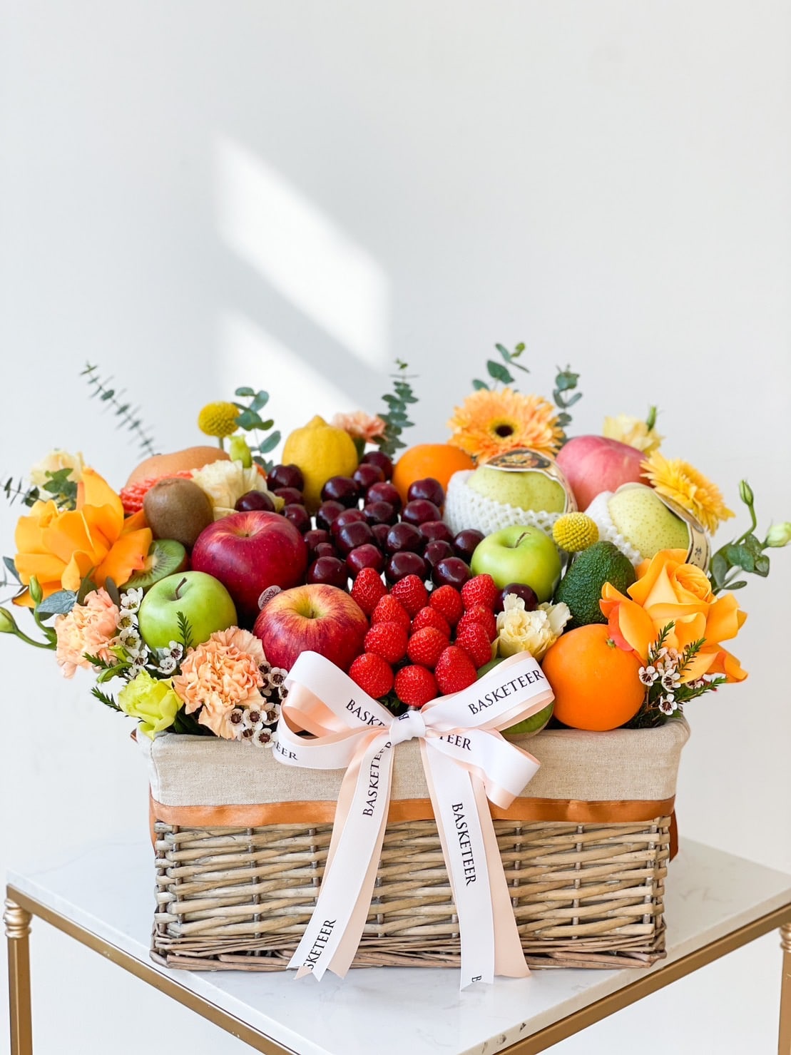A wicker basket brimming with an assortment of fresh fruits including apples, oranges, grapes, raspberries, and melon. The Blooming Fruit Gift Basket is adorned with yellow and orange flowers, with a white ribbon tied at the front inscribed with 