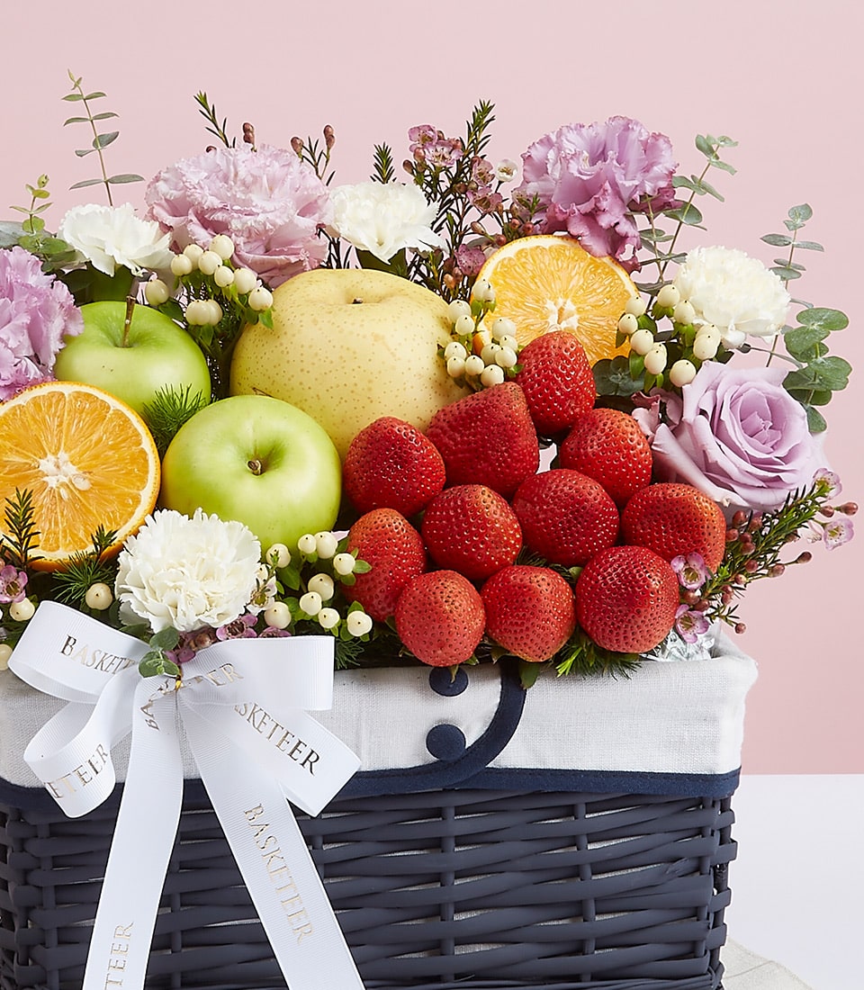The Basketeer Purple Flowers Deluxe Fruit Basket is an opulent collection of nature's finest, artfully assembled to delight the senses. Nestled within this luxurious basket are succulent fresh strawberries, crisp green apples, an exotic Asian pear, and zesty orange slices. This bountiful array is exquisitely complemented by delicate light purple roses, pristine white carnations, and vibrant sprigs of greenery. The entire ensemble is elegantly wrapped with a pristine white ribbon featuring the distinguished Basketeer logo—an invitation to indulge in pure luxury.
