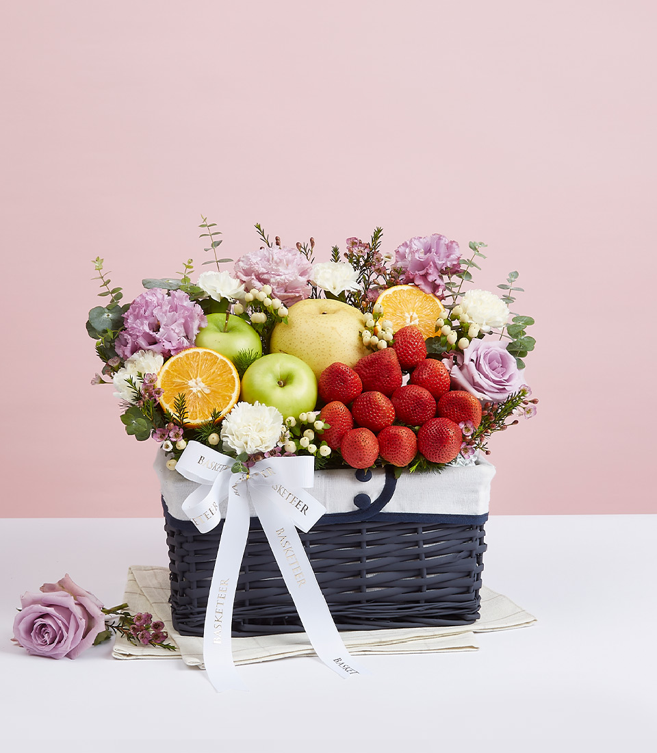 A navy-colored rattan gift basket with a beautiful fabric lining inside, filled with a variety of fresh fruits such as red strawberries, green apples, yellow and orange pears, decorated with beautiful light purple fresh flowers, and tied with a white ribbon bow with the word 