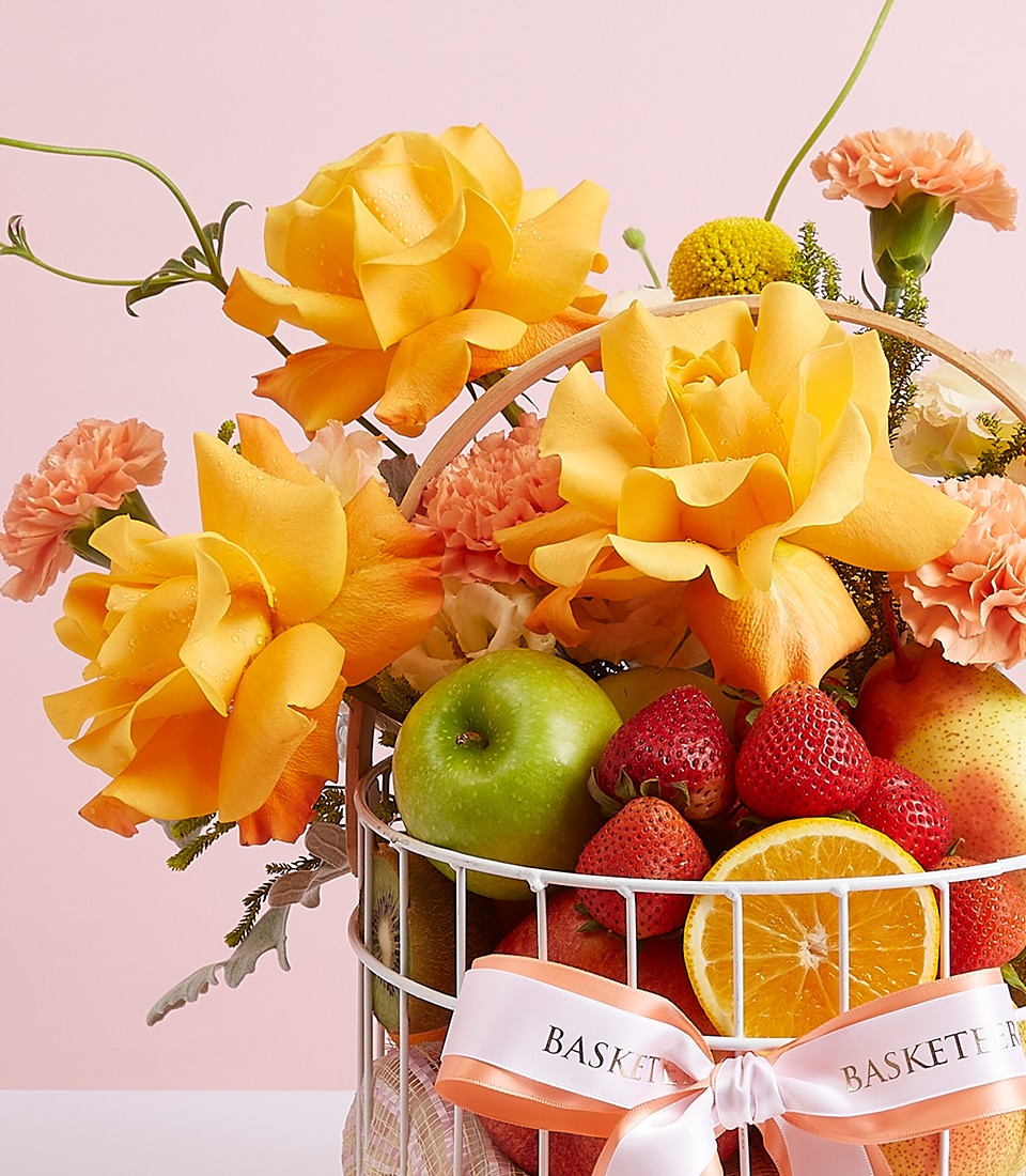 A lavish white metal basket brimming with an assortment of succulent fruits, including crisp green apples, luscious strawberries, and a perfectly halved orange. The arrangement is exquisitely enhanced with vibrant yellow roses and delicate peach carnations. The basket is elegantly tied with a pristine white ribbon that beautifully showcases the Basketeer logo and reads 
