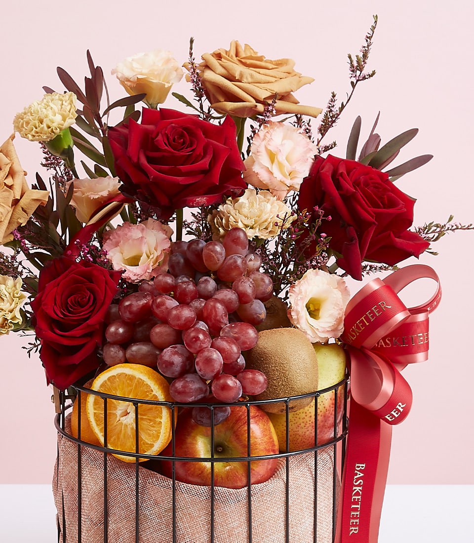 A luxurious Basketeer gift basket brimming with rich red roses, delicate pink carnations, and an assortment of vibrant, fresh fruits. Grapes cascade beside juicy oranges, crisp apples, and a tantalizing kiwi. The elegant arrangement is adorned with a stylish red ribbon proudly displaying the 
