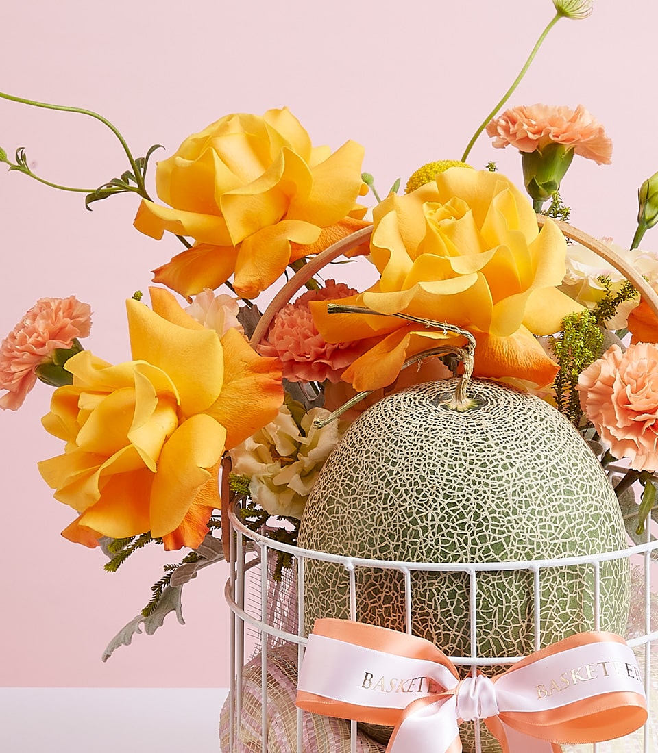A luxurious decorative arrangement showcasing a luscious melon, elegantly nestled within an exquisite white cage. The cage is adorned with vibrant yellow blossoms and delicate peach carnations for a touch of natural beauty. A pale peach ribbon, exquisitely embossed with 