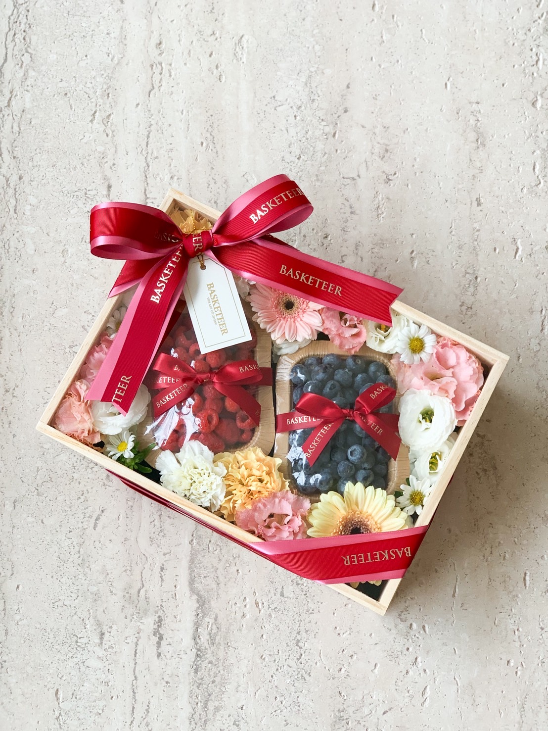 Wooden gift box with fresh raspberries, blueberries, and assorted pastel flowers wrapped with red ribbon