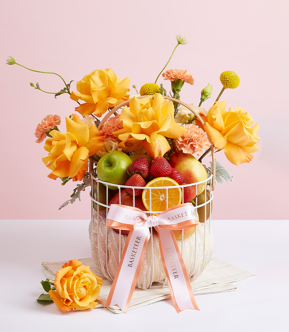 White steel frame gift basket Filled with various types of fresh fruits such as green apples. Red strawberries, red pears, and many more. Decorated with fresh flowers in yellow tones. And beautifully decorated with bows, orange and white ribbons with the word “Basketeer”