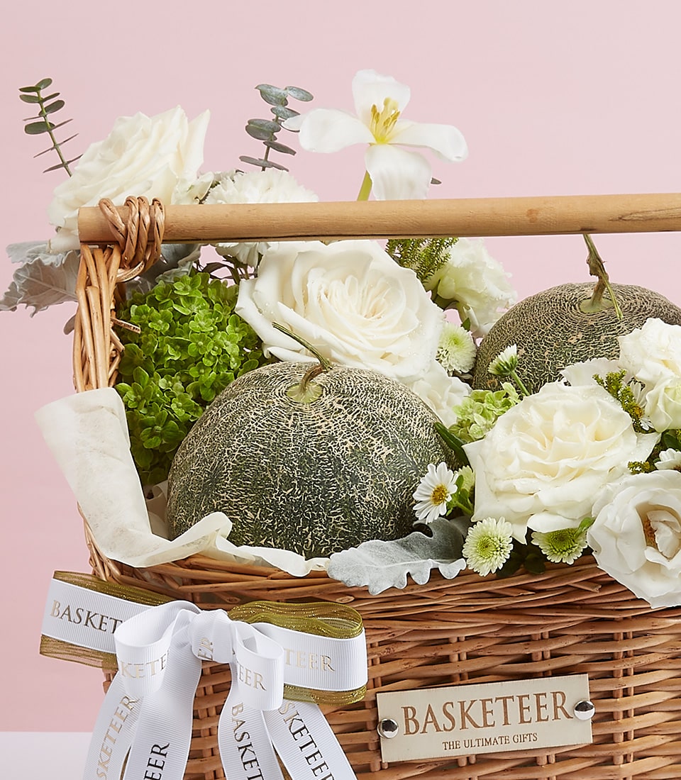 Tall brown rattan gift basket Filled with fresh fruit, 2 large melons, and white roses. Decorated with a green and white ribbon bow with the word 