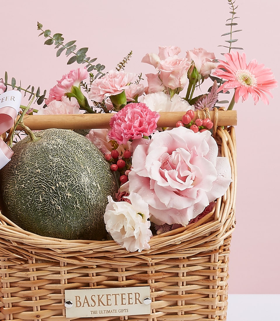 A sumptuous woven basket brimming with an exquisite assortment of blush-hued roses and delicate pink carnations, elegantly accompanied by a luscious green melon. The scene is beautifully accentuated by the 