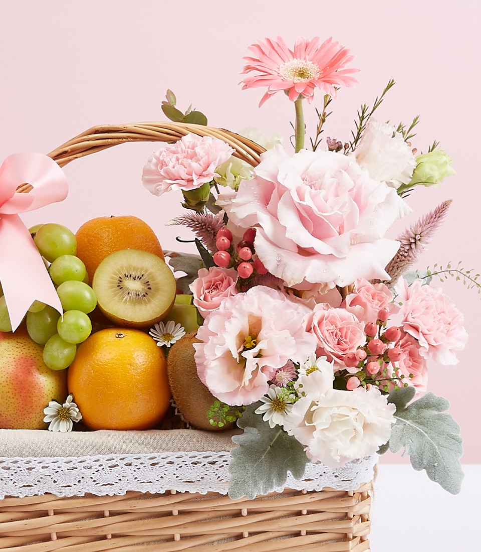 A luxurious wicker basket filled with an exquisite selection of fresh fruits, including vibrant oranges and lush green grapes, is elegantly adorned with a stunning array of pink and white flowers—roses, carnations, and daisies. A delicate pink ribbon graces the basket, featuring The Basketeer’s logo prominently. Set against a soft pink background, this scene exudes sophistication and charm.