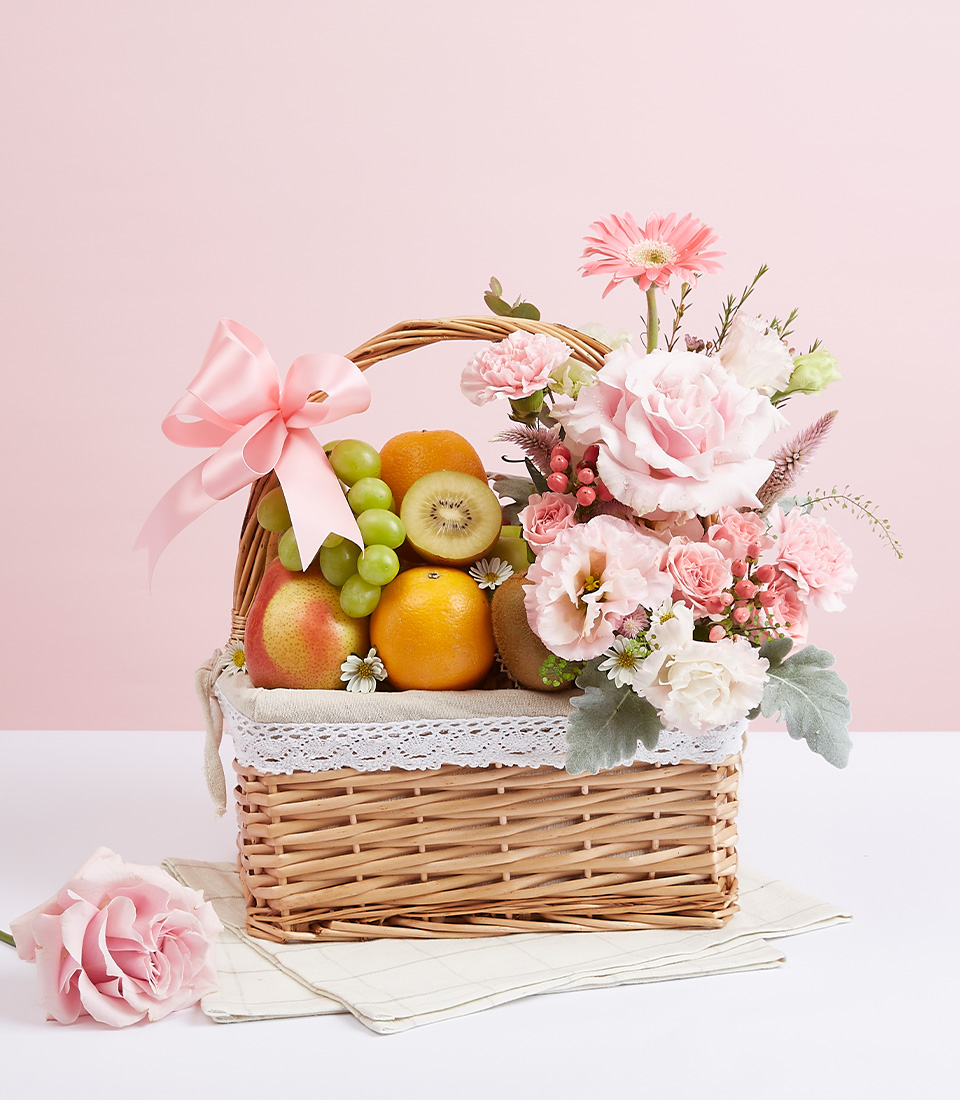 A brown rattan gift basket with fabric lining inside. Filled with a variety of fresh fruits such as red pears, green grapes, green kiwis, and many more. Decorated with fresh, sweet, beautiful light pink flowers and tied with a light pink ribbon bow.