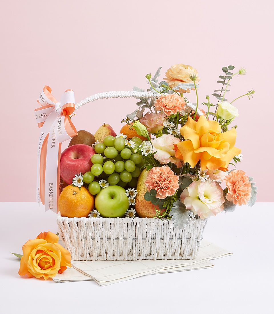 A white gift basket filled with various types of fresh fruits such as red apples, green apples, green grapes, and many more, decorated with fresh flowers in beautiful light orange tones, and adorned with light orange and white ribbons with the words 