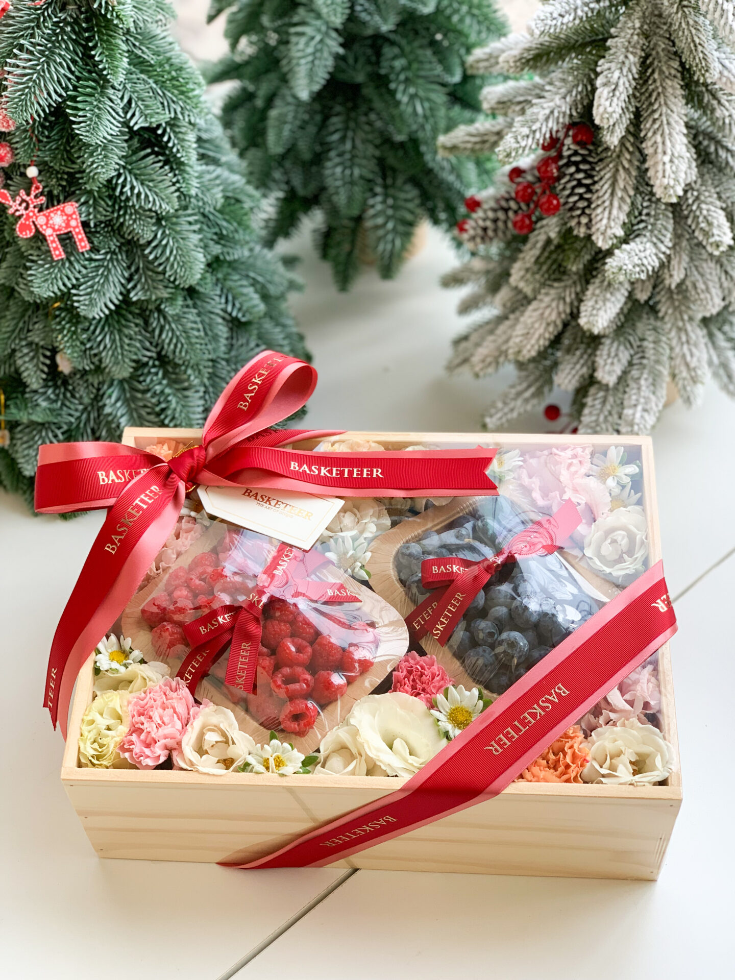 A luxury gift box featuring fresh raspberries, blueberries, and pastel flowers with a red ribbon, set against a festive background