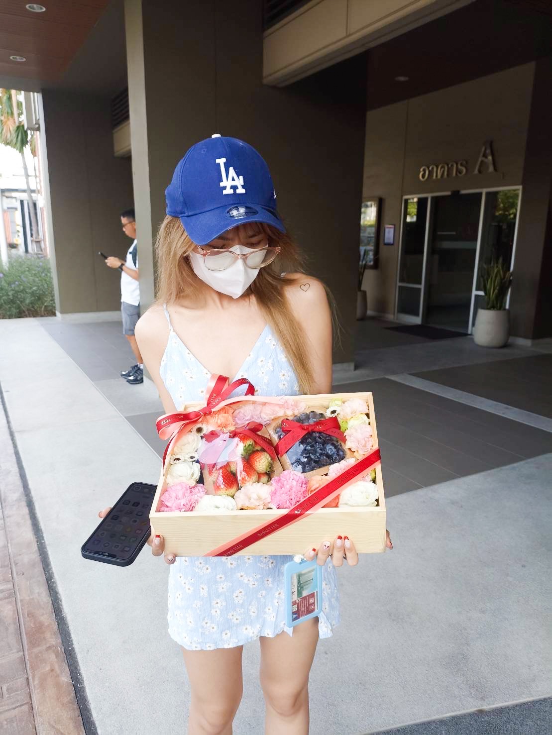 A person wearing a blue dress, blue baseball cap, and face mask holds a Sweet Berry Gift Set filled with sushi and fruit, wrapped with a red ribbon. They stand in front of a building entrance with another person in the background.