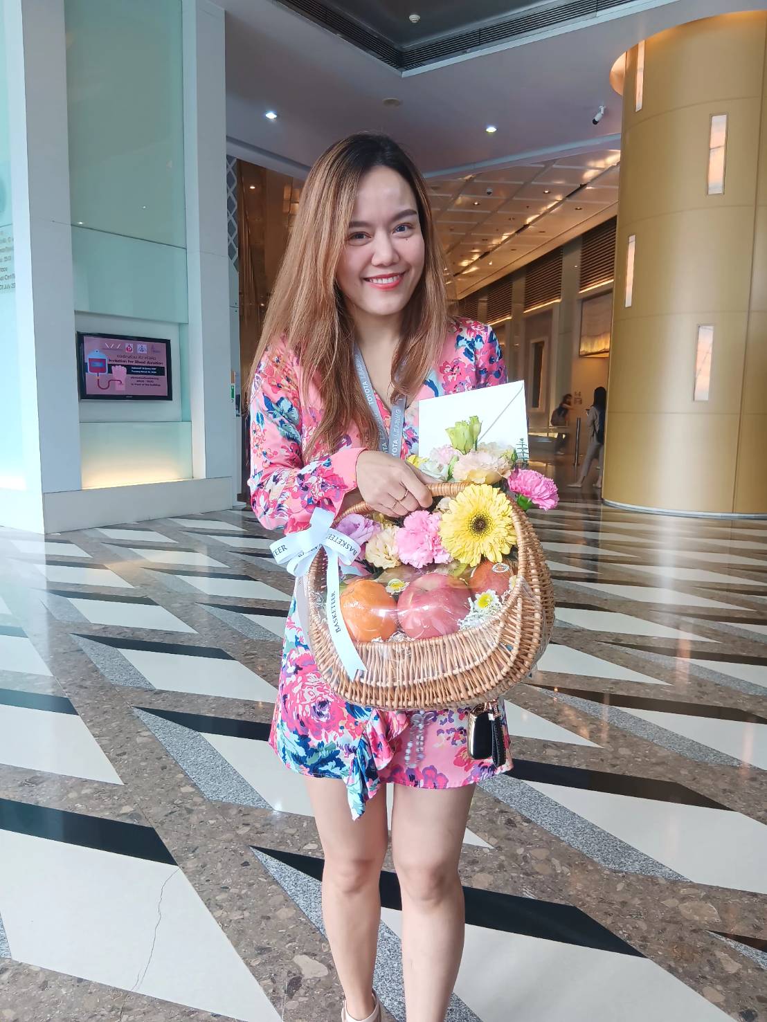 A smiling woman stands in a brightly lit building, holding a wicker basket filled with a luscious fruit harvest and flowers. She is wearing a colorful floral dress and has long hair. The floor has a geometric pattern of black and white stripes, and the background features modern decor.