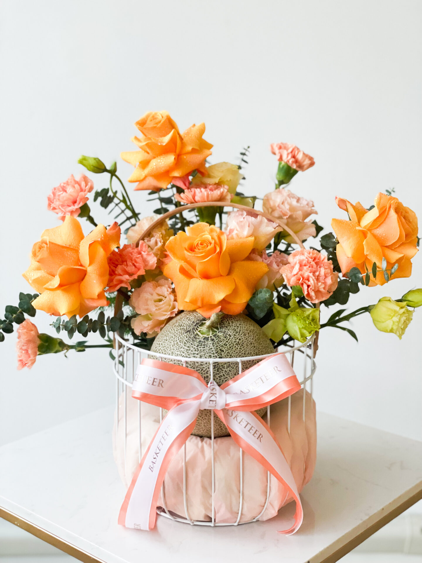 A decorative white wire basket holds a variety of orange and yellow flowers, including roses and carnations. An uncut melon rests in the center of the arrangement. The basket is adorned with a white and pink ribbon bow. The background is plain white.