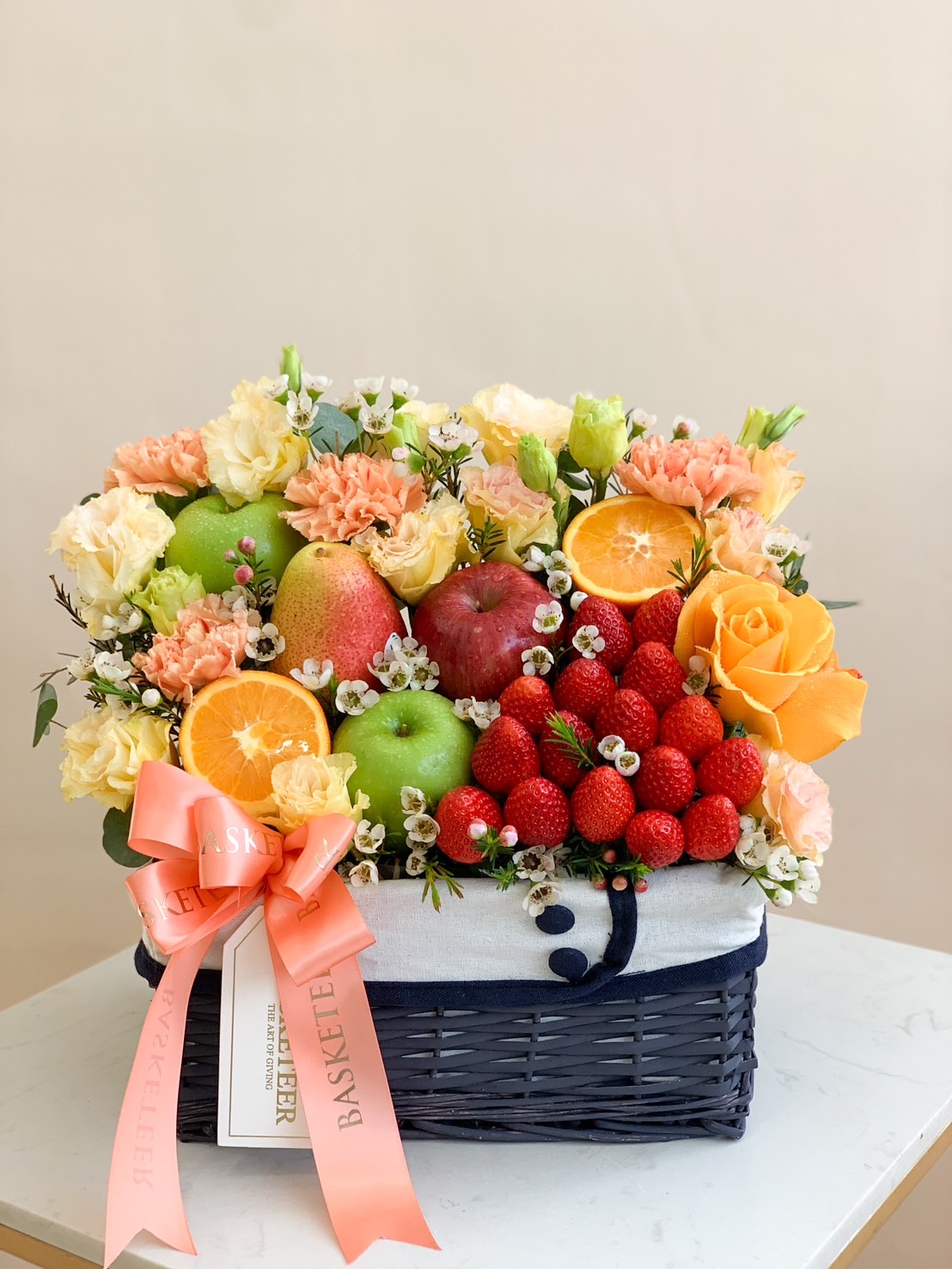 A premium fruit and floral gift basket beautifully arranged with fresh strawberries, apples, oranges, pears, and delicate blooms in shades of peach, cream, and yellow. Wrapped with an elegant coral ribbon, making it a perfect luxury gift.