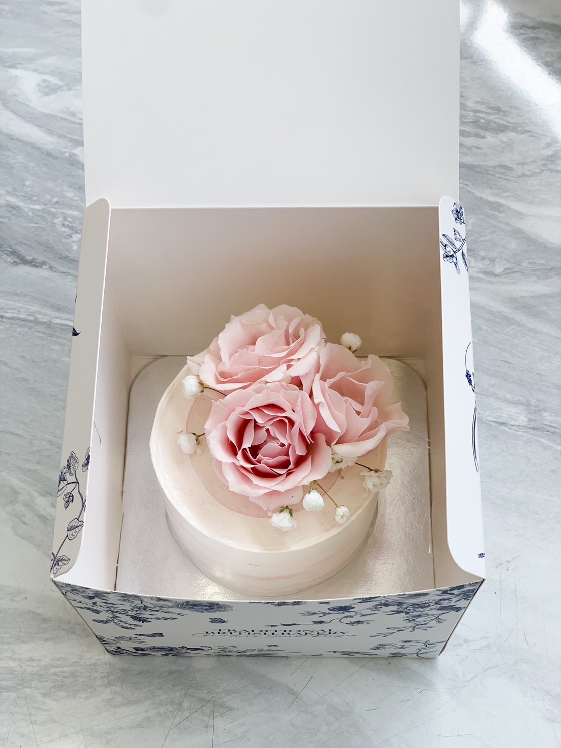 A delicate buttercream cake adorned with fresh pink roses and baby’s breath, beautifully placed in a chic floral-patterned box. Perfect for birthdays, anniversaries, and special celebrations.