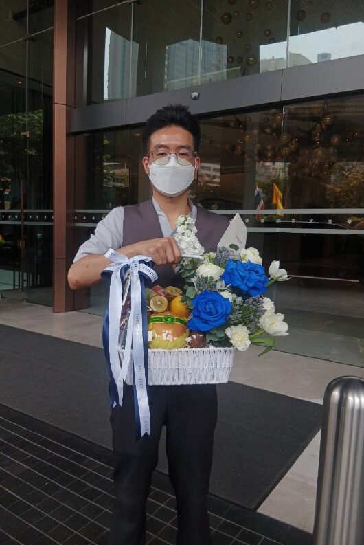 A luxurious white fruit basket filled with fresh apples, pears, grapes, and exotic fruits, complemented by blue and white floral arrangements and wrapped in a stylish ribbon.
