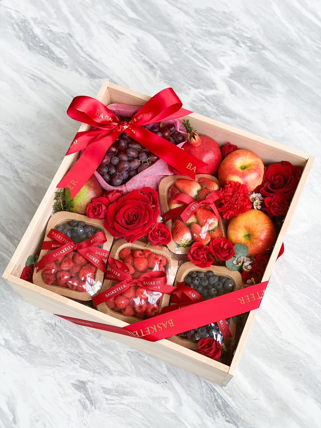A wooden box filled with various fresh fruits including apples, grapes, strawberries, and blueberries, dubbed the Mixed Berry Joy Gift. The box is decorated with red roses and tied with red ribbons. The delightful arrangement sits on a marble surface.
