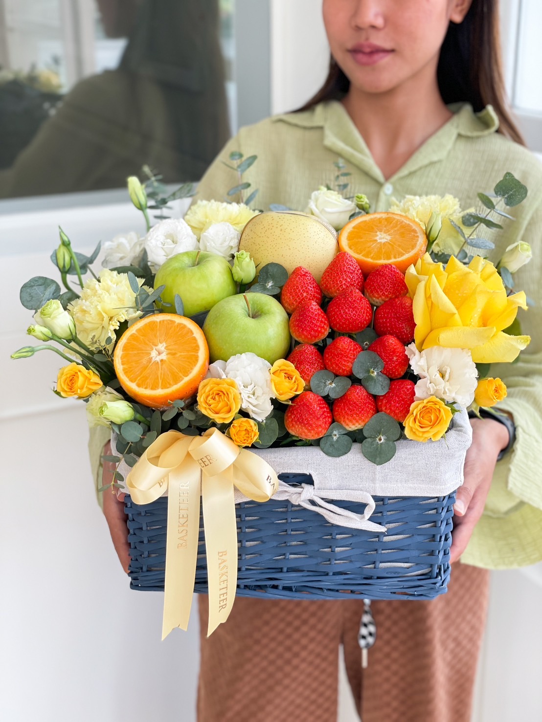 A person holding a blue wicker basket filled with a variety of fresh fruits, including strawberries, apples, oranges, and a pear. The arrangement is accented with yellow, white, and purple flowers and greenery, complete with a cream-colored ribbon emblazoned with 