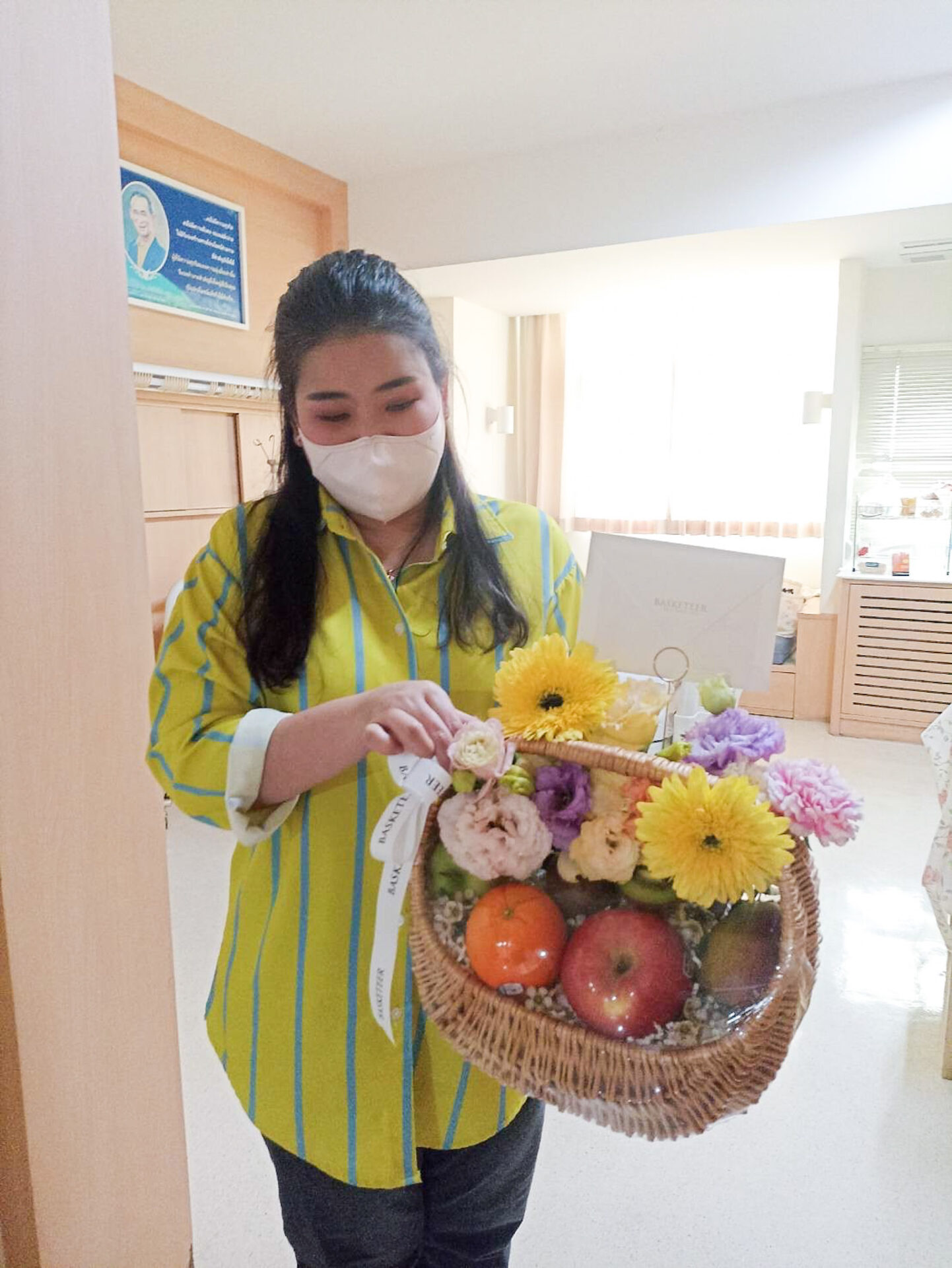 A woman wearing a yellow and green striped shirt and a face mask holds a wicker Luscious Fruit Harvest Basket filled with yellow and purple flowers, apples, and oranges. She stands in a bright, well-lit room with a framed portrait on the wall and a desk in the background.