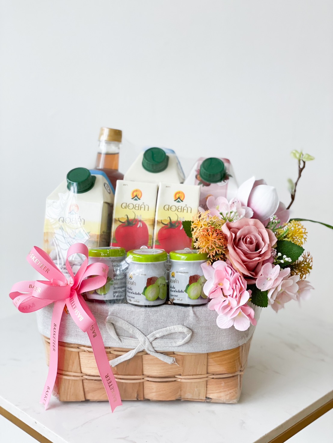 A wooden basket filled with various drinks and decorated with a pink ribbon and bow. It contains cartons of fruit juice, several jars, and a bottle. The basket is adorned with artificial flowers including roses and other blossoms against a simple white backdrop.
