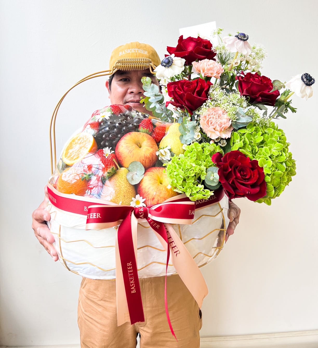 A person in a cap and pink shirt holds a large Harvest Fruit Floral Basket, filled with assorted fruits and a bouquet of red, pink, and white flowers. The basket is wrapped in plastic and adorned with red ribbons labeled 