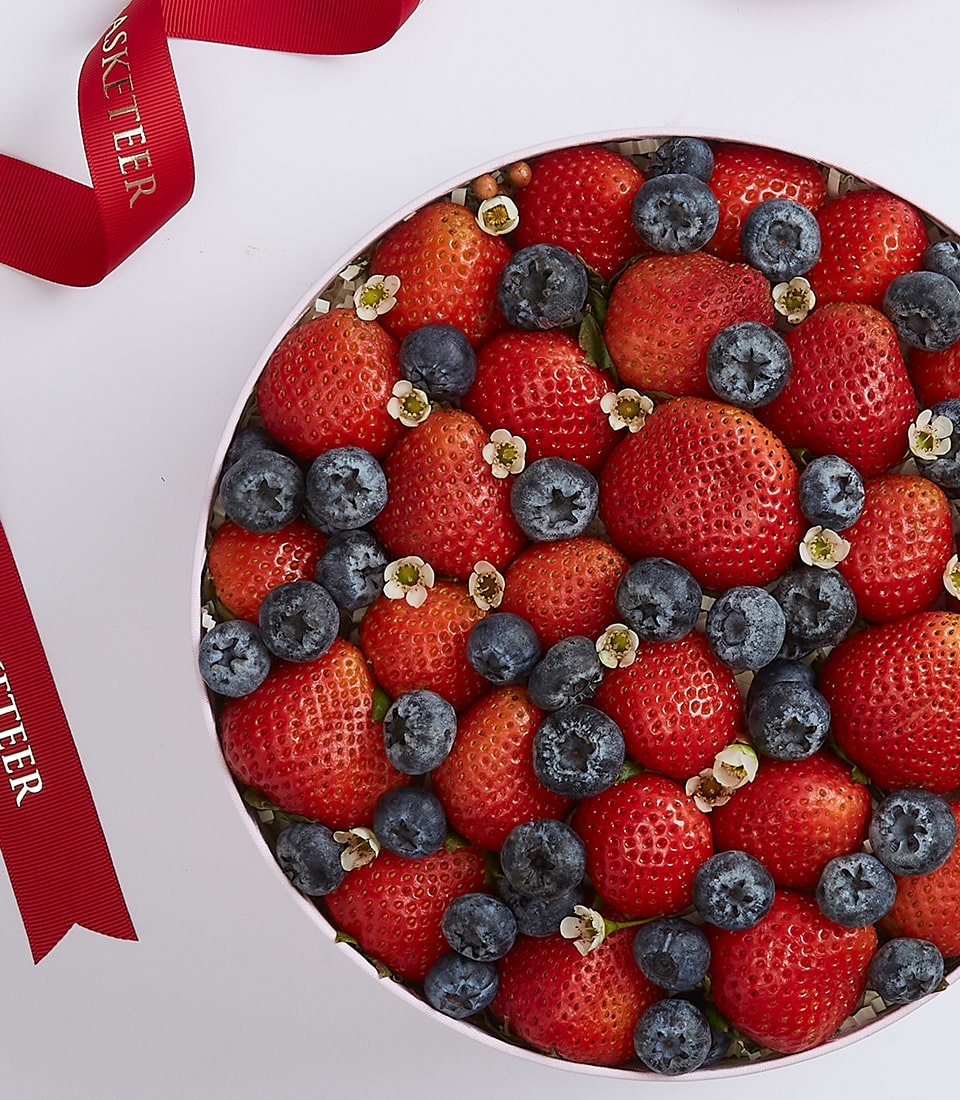 A sumptuous round fruit tart crowned with a bountiful array of fresh, plump strawberries and luscious blueberries, elegantly garnished with delicate white flowers. Red ribbons embossed with 