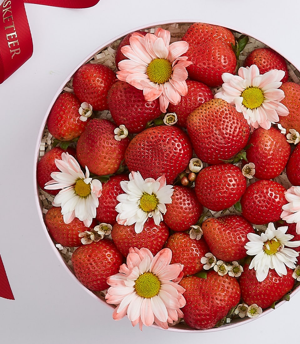 A round box filled with fresh strawberries and decorated with white and pink daisies as well as small white flowers. A red ribbon with some printed text is partially visible on the top left corner.