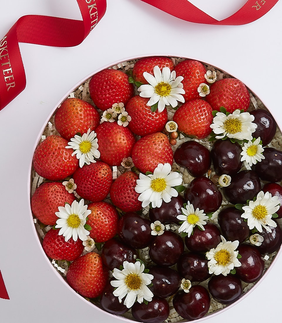 A deluxe Basketeer Sweet Berry Gift Box showcases a beautifully arranged round basket overflowing with plump, fresh strawberries and succulent dark cherries, elegantly interspersed with delicate white daisies. The basket rests gracefully on a pristine white surface, accented by luxurious red ribbon pieces bearing the distinguished Basketeer logo.