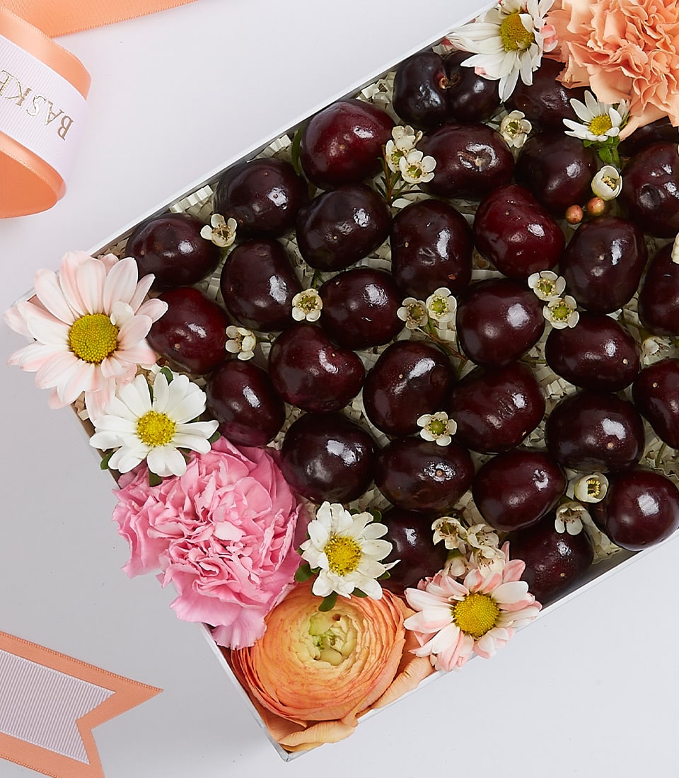 A lavish Basketeer Cherry Bliss Gift Box overflowing with luscious dark cherries, adorned with an exquisite arrangement of pristine white daisies, delicate pink carnations, and enchanting peach ranunculus. The presentation is artfully completed by an elegant Basketeer-branded ribbon that adds a touch of sophistication. This luxurious ensemble rests gracefully against a pristine white backdrop, inviting you to indulge in its opulence.