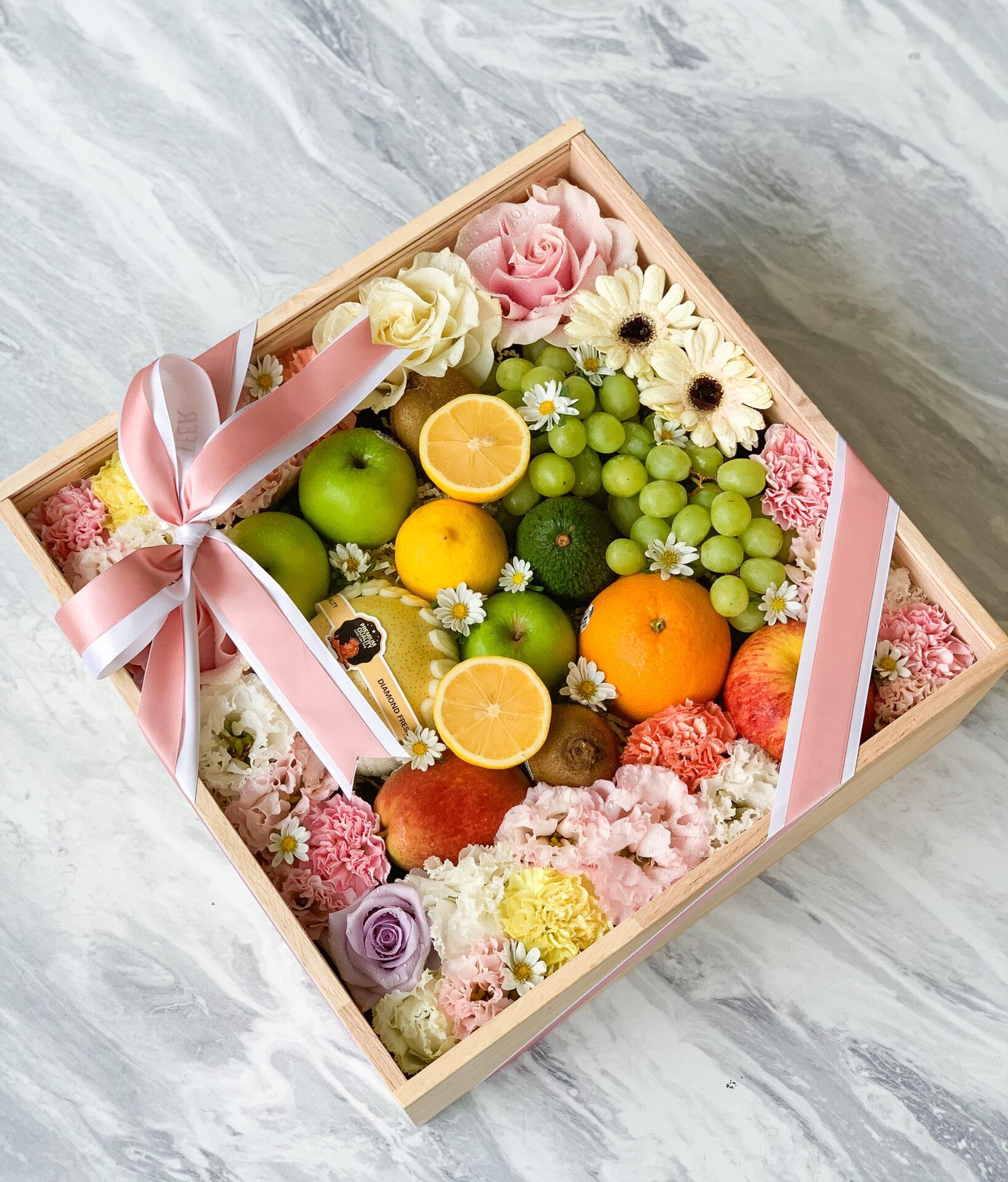A special Fresh Fruit Box filled with a variety of fruits and flowers. The arrangement includes apples, oranges, grapes, lemons, and avocados, surrounded by roses, daisies, carnations, and other blooms. A pink ribbon with a bow is tied around the box.