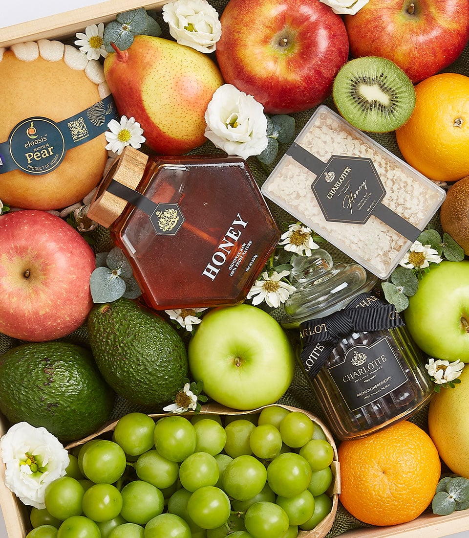 The opulent Splendor Fruit Gift Box by Basketeer showcases an elegant wooden tray filled with a premium selection of fresh fruits—crisp apples, juicy kiwi, succulent pears, creamy avocados, plump grapes, and vibrant oranges. Enhancing this luxurious ensemble are a jar of pure honey, a delightful jar of charlotte preserves, an exquisite box of gourmet chocolates adorned with the iconic Basketeer ribbon. Delicately arranged small white flowers add a touch of sophistication to this regal display.