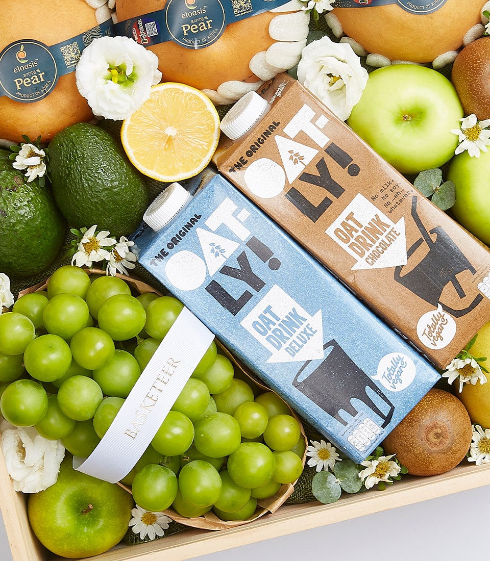 Indulge in the opulence of the Deluxe Harvested Bounty Box by Basketeer. Nestled within a sophisticated wooden crate, you'll discover an exquisite selection of lush green grapes, crisp apples, creamy avocados, and a vibrant halved lemon. Elevate your experience with two cartons of Oatly oat drink—one offering the pure essence of original flavor and the other presenting a decadent chocolate delight. Accentuated with delicate small white flowers and finished with an elegant ribbon proudly emblazoned with 