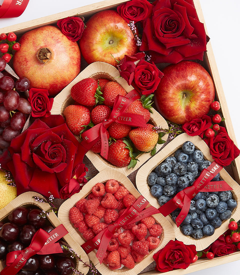 A luxurious wooden box brimming with a vibrant array of fresh red and purple fruits, including succulent pomegranates, crisp apples, juicy strawberries, luscious grapes, tangy raspberries, sweet blueberries, and plump cherries. Elegantly adorned with fragrant red roses and lavishly arranged with opulent red ribbons proudly bearing the Basketeer insignia. This 